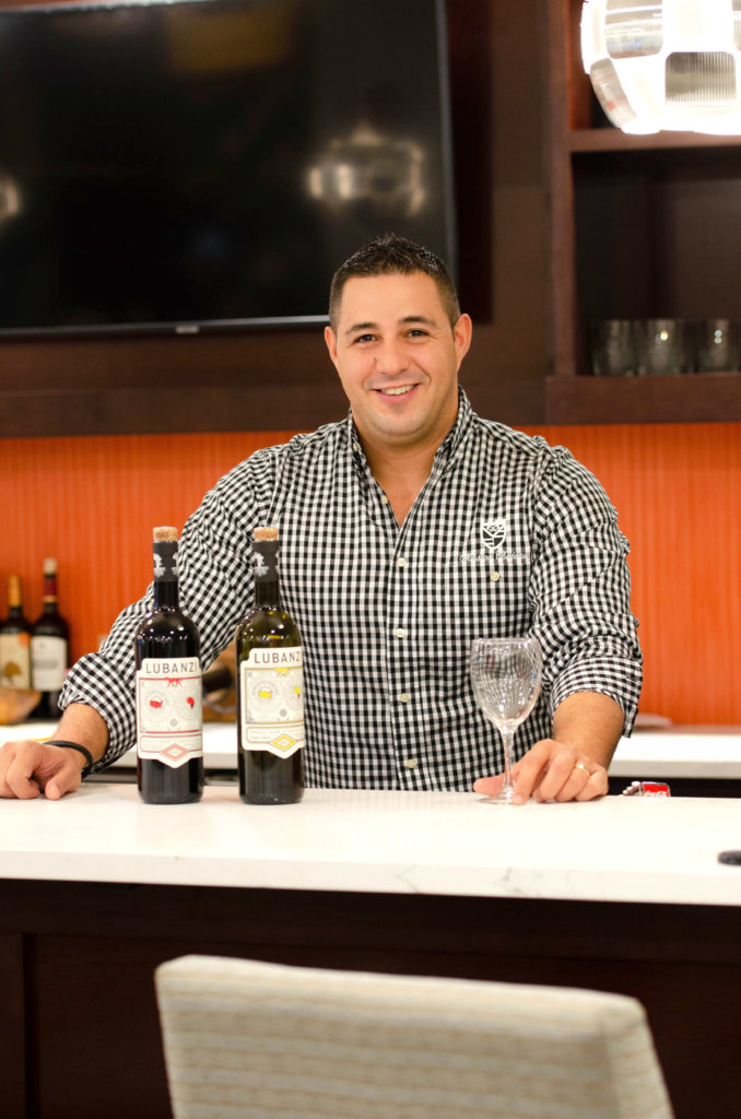 A young male bartender at the bar with an empty glass of wine and wine in full bottles next to him.