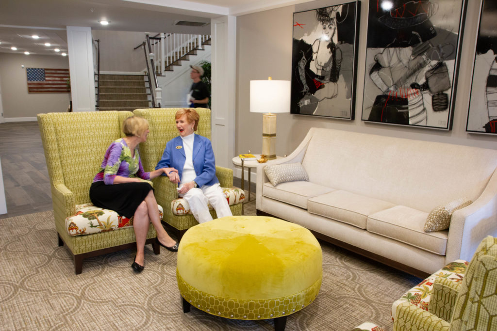 Two female residents chatting together on a yellow couch in a living room with two couches and a foot rest.