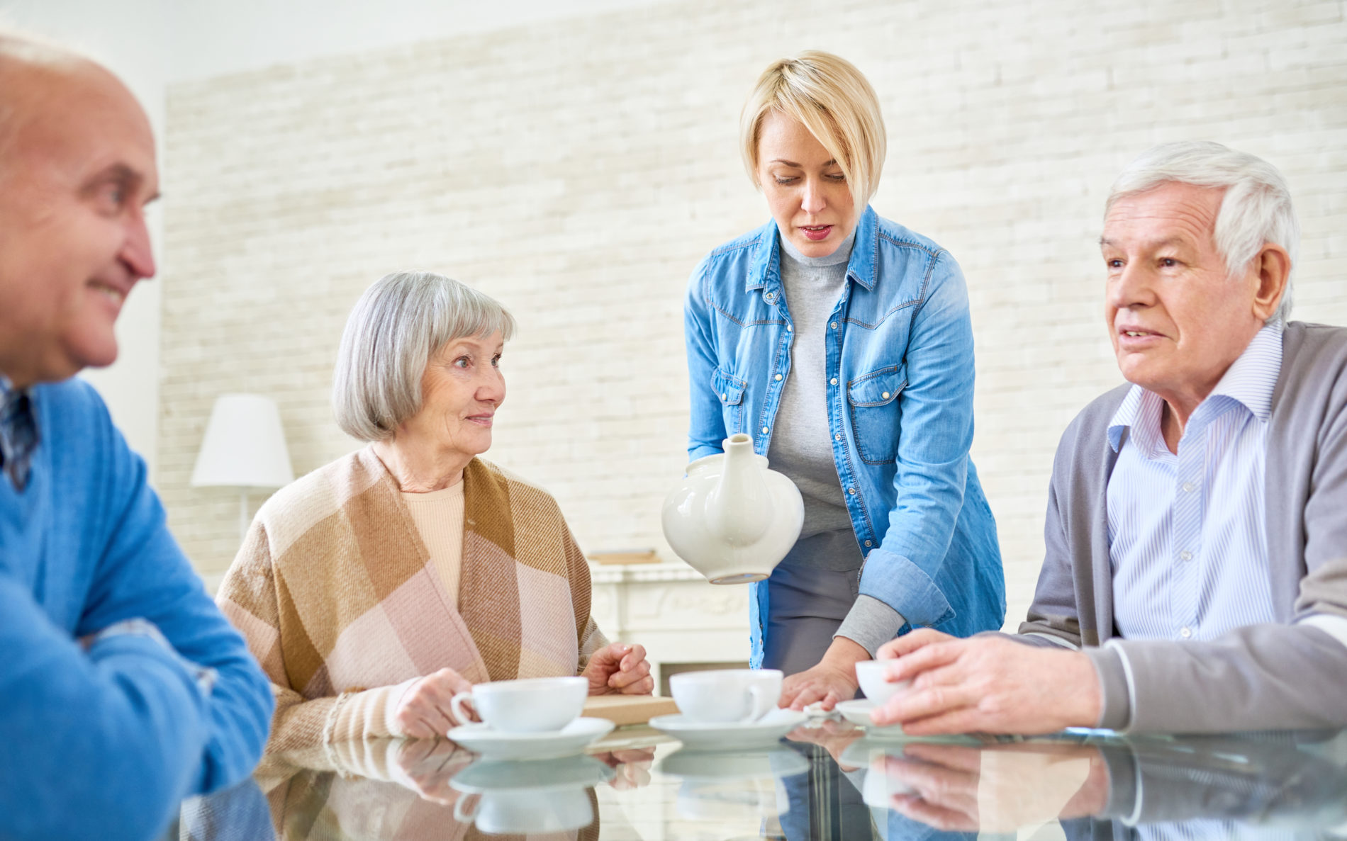 Woman pouring tea for senior patients in assisted living home sitting at table and chatting.