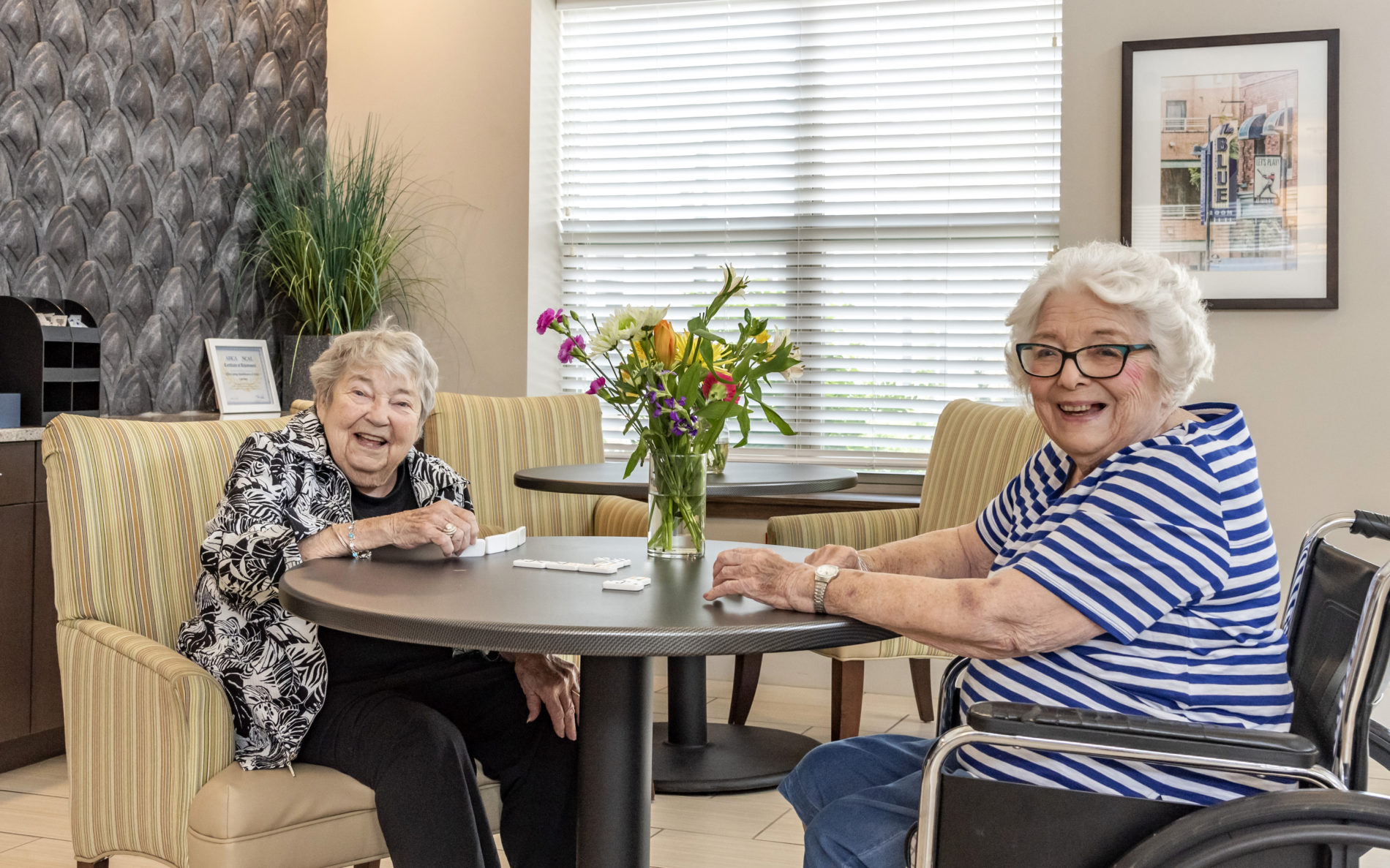 Two elderly females smiling and playing dominoes at table with flowers